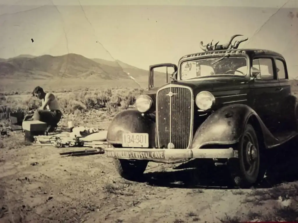 A vintage black car with a license plate reading 134-901 is parked on a dusty road. A man sits on the ground to the left, surrounded by miscellaneous items. Mountains rise in the background under a clear sky.