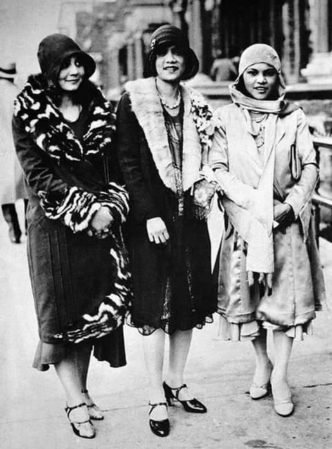 Three women in 1920s fashion stand on a city street. They wear dresses with fur coats and cloche hats, embodying the style of the era. The woman on the left wears a patterned coat, and the other two sport lighter dresses with shawls.