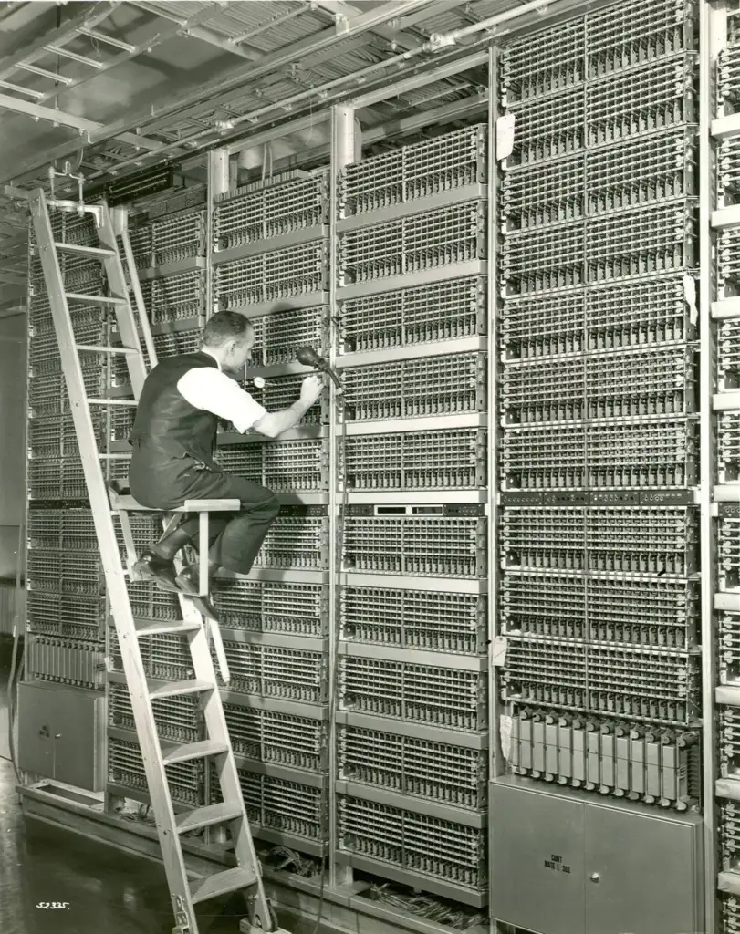 A man in a vest and tie is sitting on a ladder, working on a large, intricate wall of vintage electronic equipment with numerous wires and components in a room with metallic hardware.