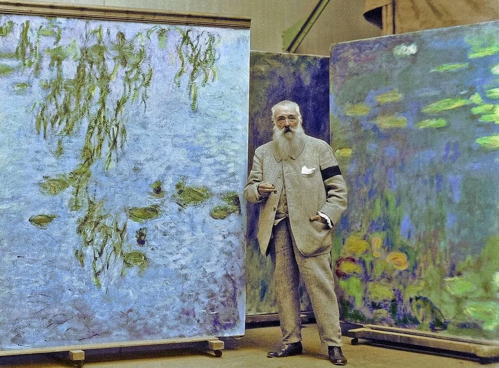 An elderly man in a suit stands in a studio, surrounded by large impressionist paintings of water lilies and willow trees. The paintings are rich in blues and greens, displaying an aquatic theme.