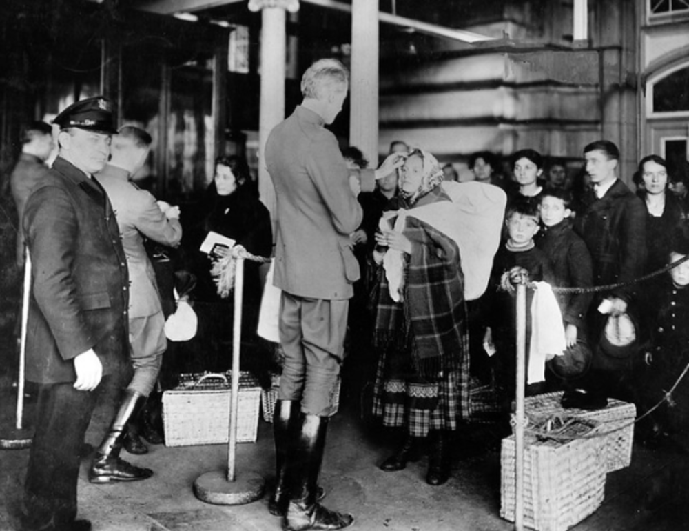 A black and white historical photo shows a group of immigrants at a customs checkpoint, possibly Ellis Island. An officer is inspecting a woman holding a baby. Others wait with baskets and luggage amidst a busy atmosphere.