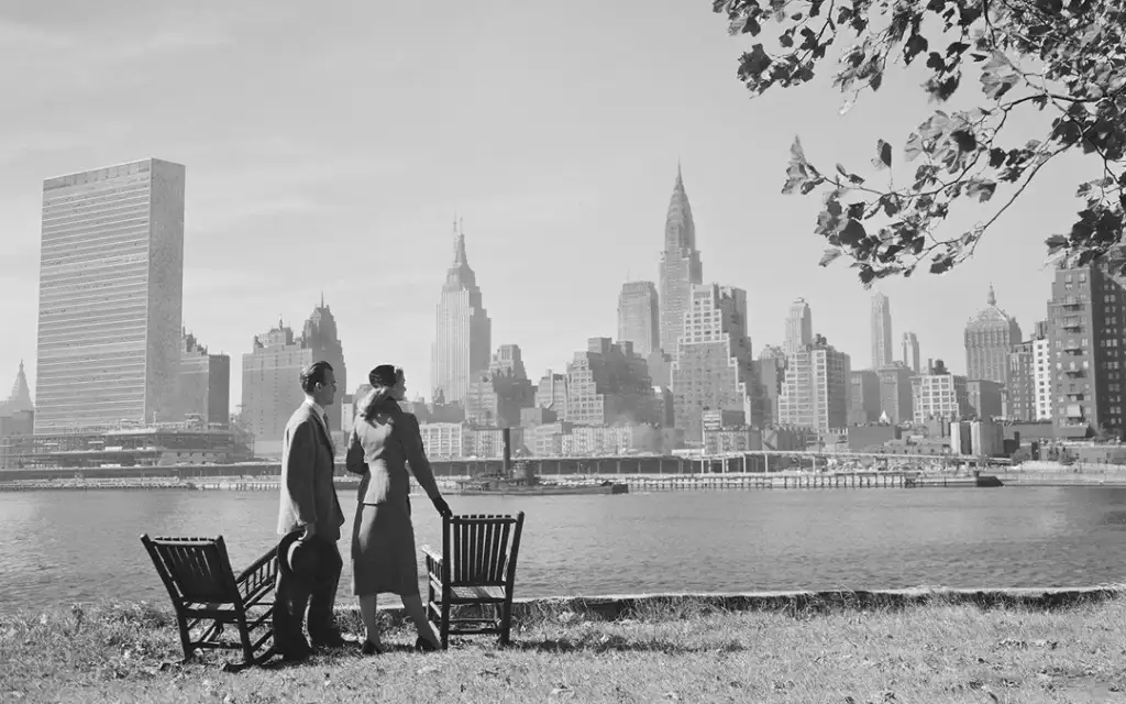 A couple in vintage clothing stands at the edge of a grassy area, overlooking the water. The New York City skyline, featuring mid-20th century skyscrapers, including the Chrysler Building, is visible in the background. Two empty chairs are nearby.