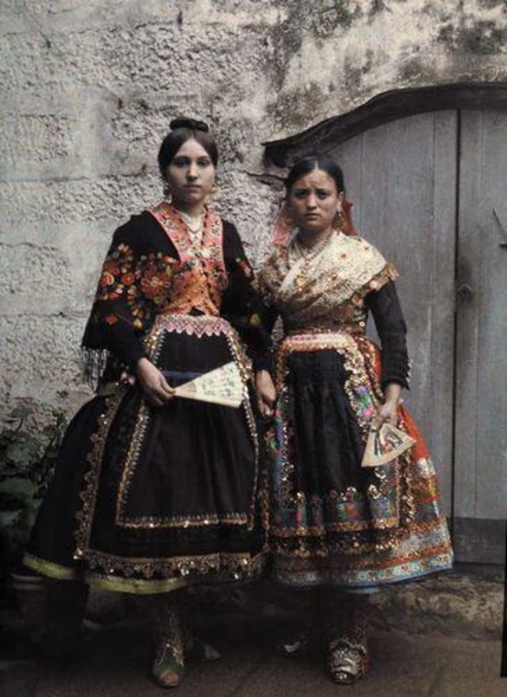 Two women in ornate traditional dresses stand in front of a rustic wooden door. Their attire features intricate embroidery and patterns. Each holds a decorative fan. The setting suggests an old-world or cultural atmosphere.