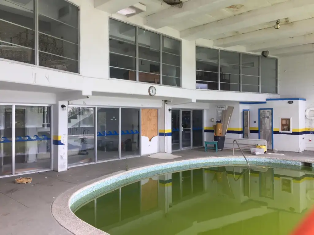 Abandoned indoor swimming pool with murky green water, surrounded by glass doors and windows. The walls are white with blue and yellow stripes. A clock is mounted above the entrance. The space appears neglected and in disrepair.