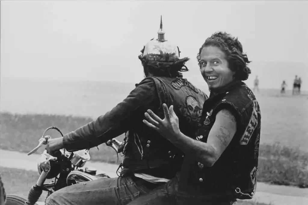 Two people ride a motorcycle. The front rider wears a helmet with a spike and has a dark vest. The back rider is looking back, smiling with an exaggerated expression, wearing a similar vest. The background shows a grassy area and people in the distance.