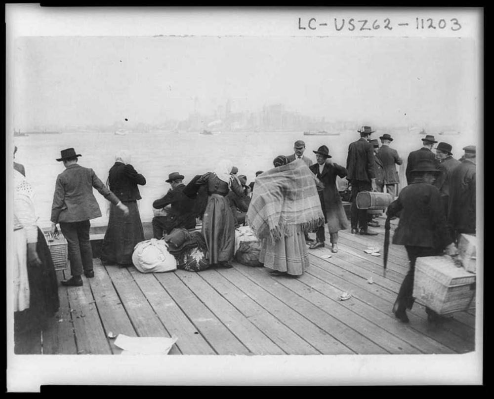 A black and white photo depicts a group of people, possibly immigrants, gathered on a wooden dock with luggage and bundles. They are dressed in early 20th-century clothing, and the skyline of New York City is faintly visible in the background.