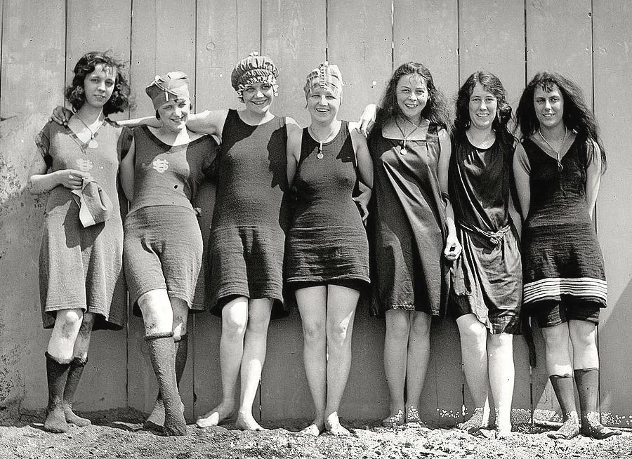 Seven women in vintage swimsuits pose cheerfully by a wooden wall. They wear various types of swim caps and have arms linked, smiling at the camera. The black-and-white image suggests a historical setting.