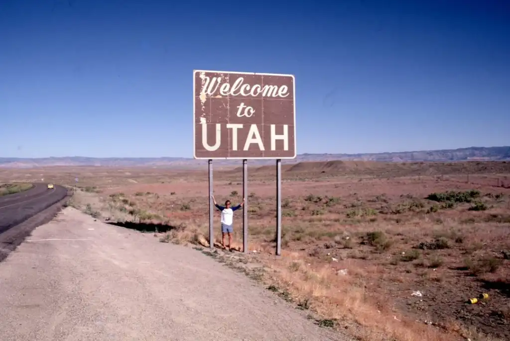 A person stands beneath a large sign that reads "Welcome to Utah" on a desert road. The landscape is barren with sparse vegetation, under a clear blue sky.