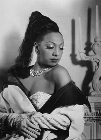 A woman in an elegant gown sits with a composed expression, looking down to her side. She has a high ponytail and wears a sparkling necklace and earrings. The background features a candelabra. Black and white photo.