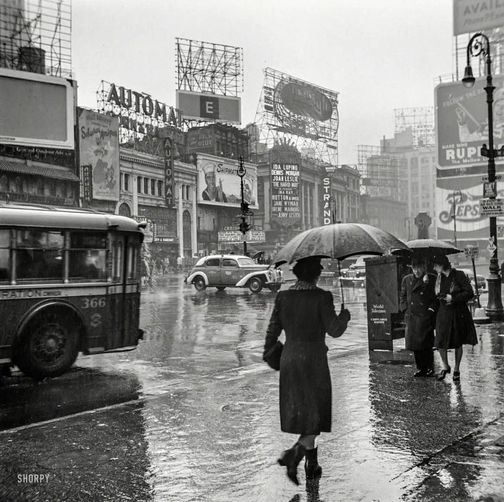 A black and white photo of a rainy urban street scene in the 1940s. A woman with an umbrella walks on a wet sidewalk, while cars, buses, and people move around. Billboard signs and storefronts are visible in the background.