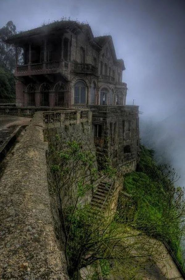 A mysterious, abandoned mansion perches on the edge of a foggy cliff, surrounded by overgrown vegetation. The stone structure features arched windows and balconies, with a misty landscape stretching into the distance on the right.