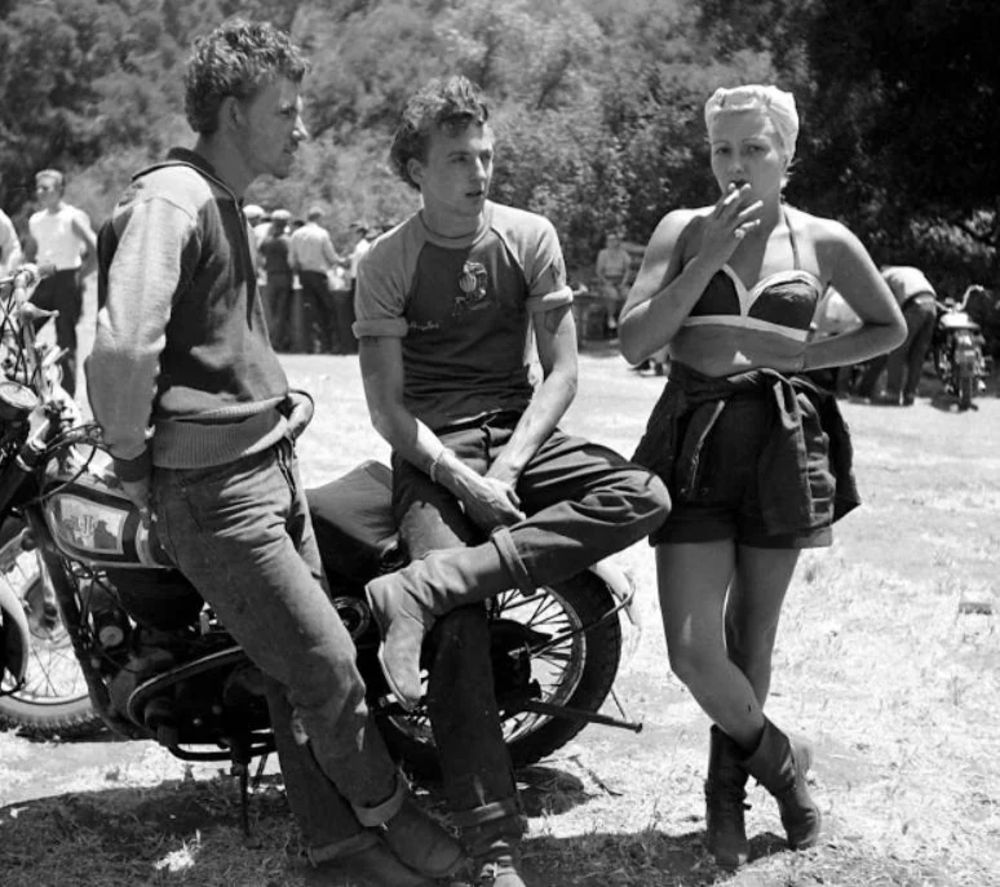 Three people in a vintage setting: two sit on motorcycles while a woman in a bikini top and shorts stands smoking. The scene is outdoors with bikes and people in the background, suggesting a biker gathering.