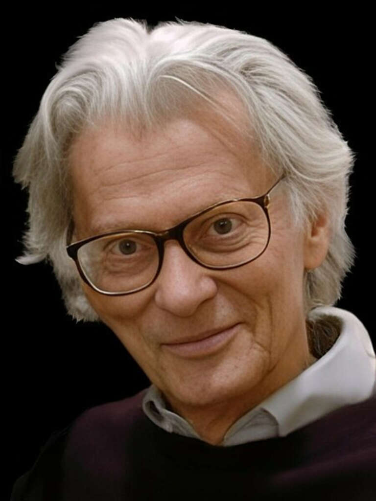 An older man with white hair and glasses smiles slightly against a black background. He wears a dark sweater over a light collared shirt.