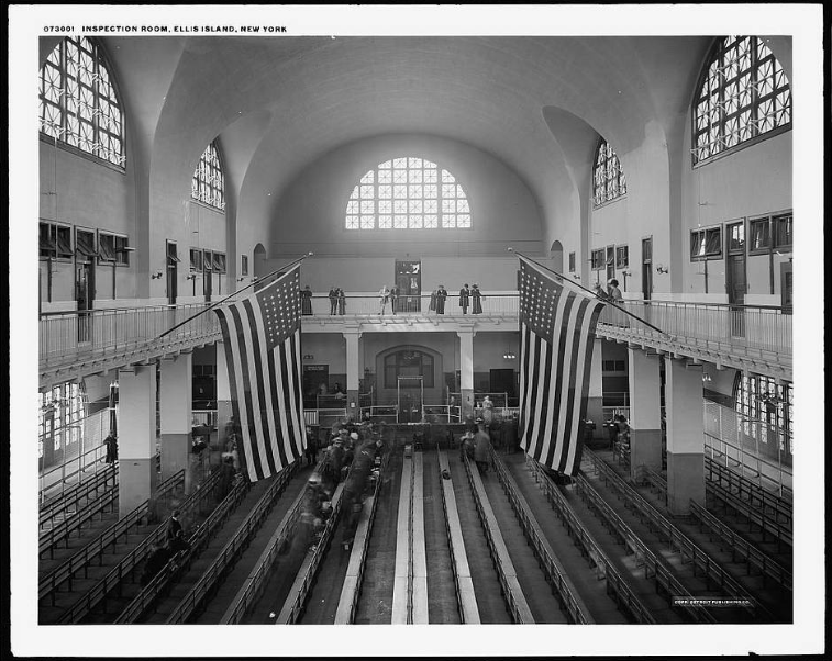 Black and white photograph of the inspection room at Ellis Island, New York, featuring two large American flags hanging from the balcony. Arched windows provide natural light. People are gathered on the main floor and upper balcony.