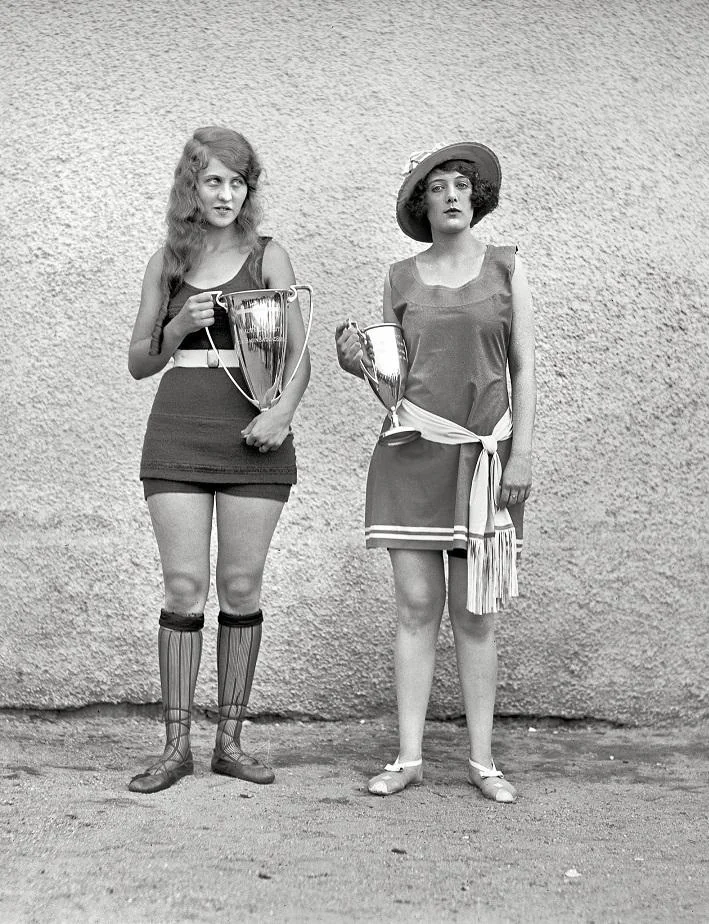 Two women in vintage swimsuits holding large trophies. The woman on the left wears knee-high socks and has long, wavy hair. The woman on the right wears a hat and a sash, standing against a textured wall on a sandy surface.