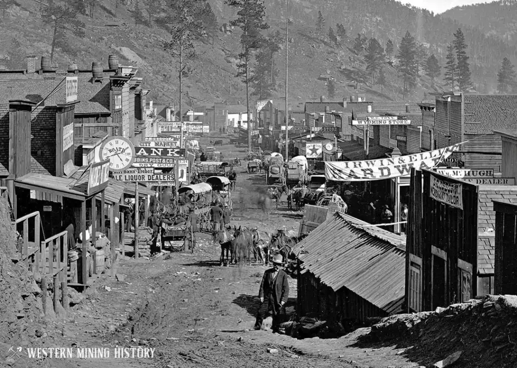 Historic black and white photo of a bustling 19th-century mining town street lined with wooden buildings. Signs advertise a bank, hardware store, and liquor dealer. Horse-drawn wagons and people populate the dirt road.