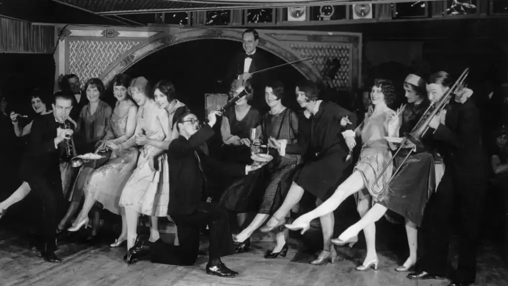 Black and white photo of a group of people from the 1920s, dressed in flapper and suit attire. They are dancing in a line, energetically kicking their legs. A band plays in the background. The scene is lively and joyful.