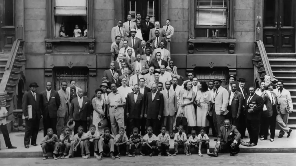 A large group of men and a few women, dressed in suits and formal wear, pose on the steps and sidewalk in front of a brownstone building. Children sit on the curb at the front. People also appear in the windows and doorways of the building.