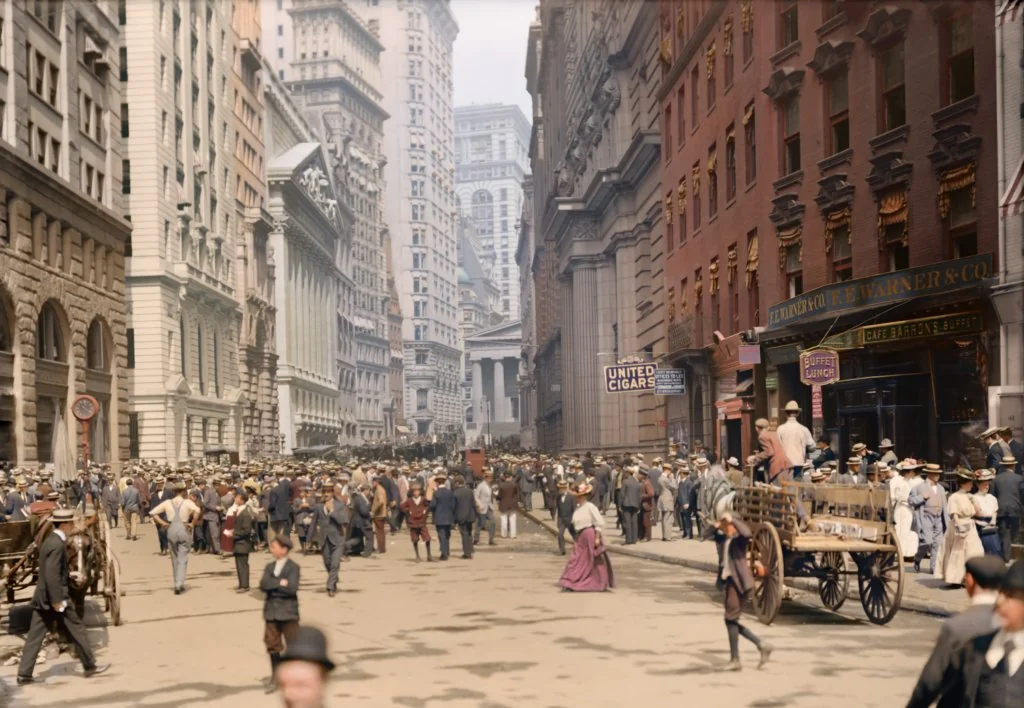 A bustling early 20th-century city street scene with crowds of people in period attire. Horse-drawn carts and vintage signs line the street, with tall buildings and a prominent columned structure in the background.