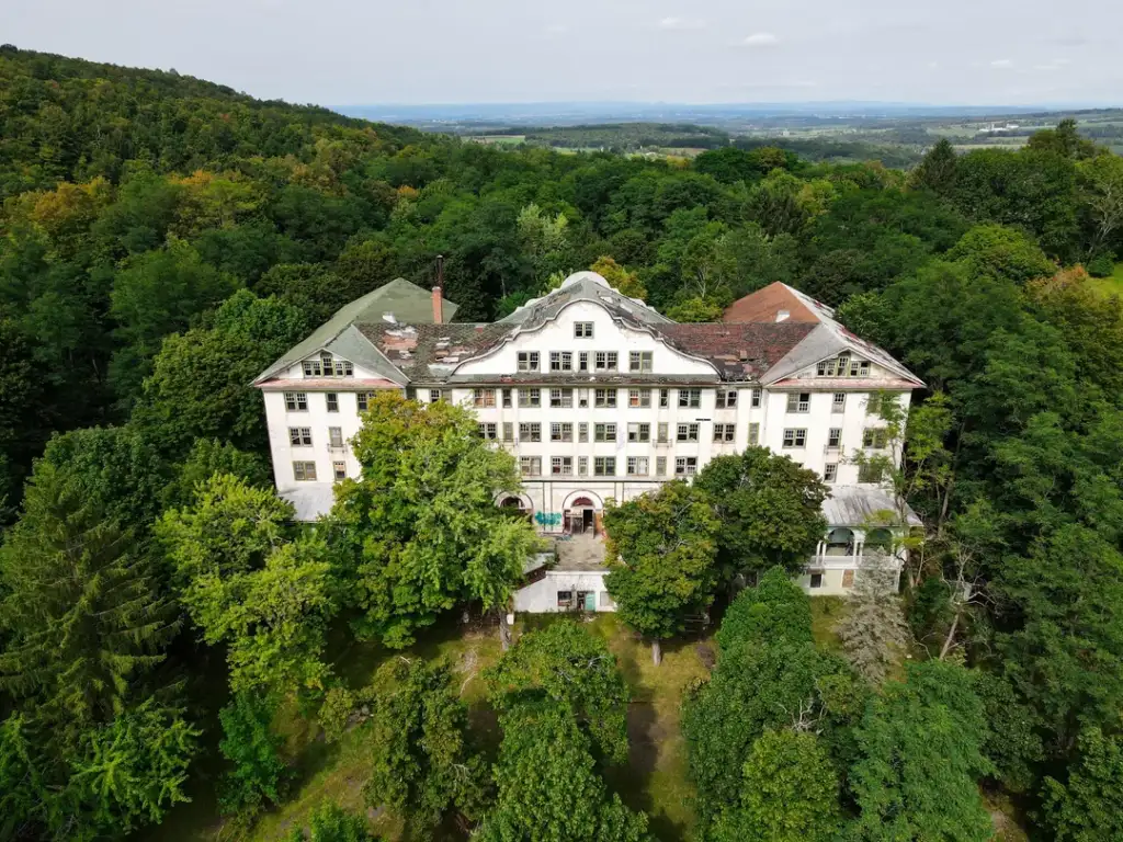 A large, old, multi-story mansion surrounded by lush green trees, viewed from above. The building has a partially damaged roof and several windows. It is set against a backdrop of rolling hills and a distant horizon.