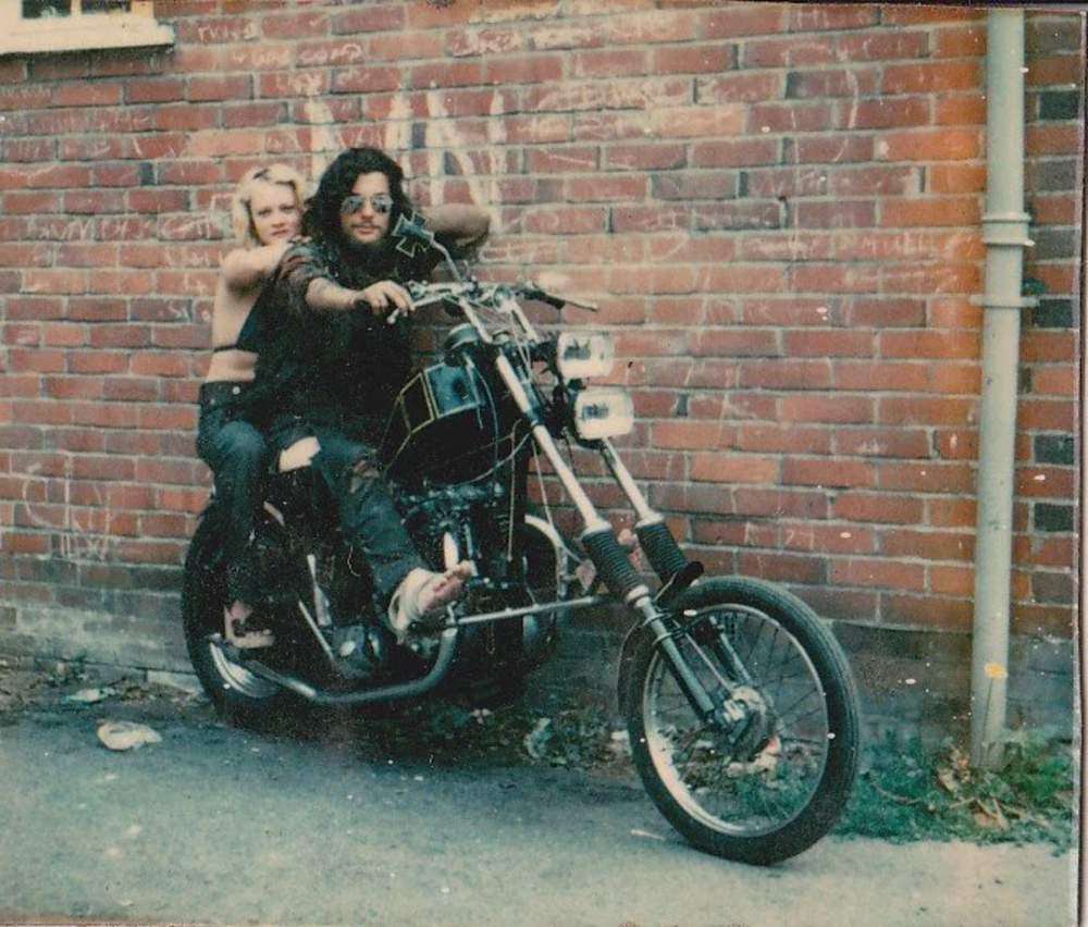 Two people sit on a motorcycle in front of a brick wall. The person in front wears sunglasses and is holding the handlebars, while the other person is behind, arms around the first. Both are casually dressed. The scene has a vintage feel.