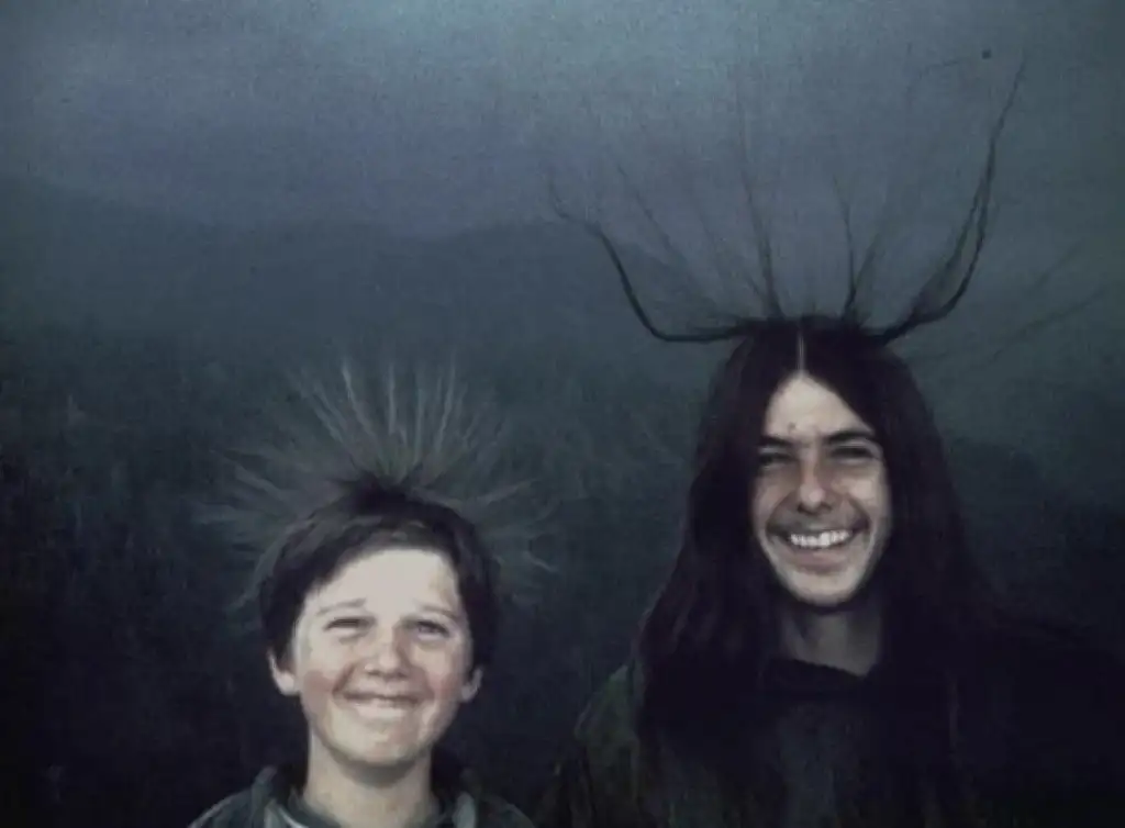 Two people outdoors with trees in the background. Both have hair standing on end, creating a spiky effect, and are smiling at the camera. The sky is overcast behind them.