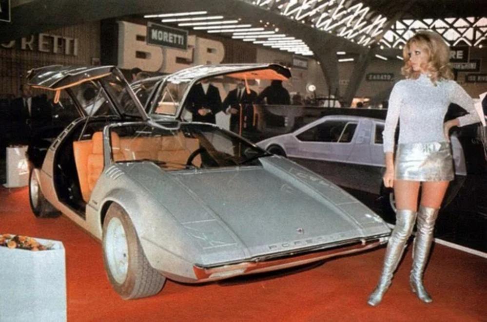 A vintage car with gullwing doors open is displayed indoors on a red carpet. A person stands beside it, wearing a silver top, a mini skirt, and knee-high boots. The setting appears to be an auto show or exhibition.