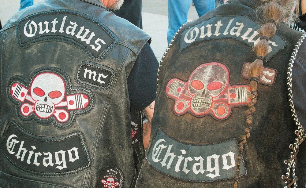 Two people wearing leather vests with "Outlaws mc Chicago" patches and a skull emblem. One person has a long braided ponytail. The vests appear rugged and worn.