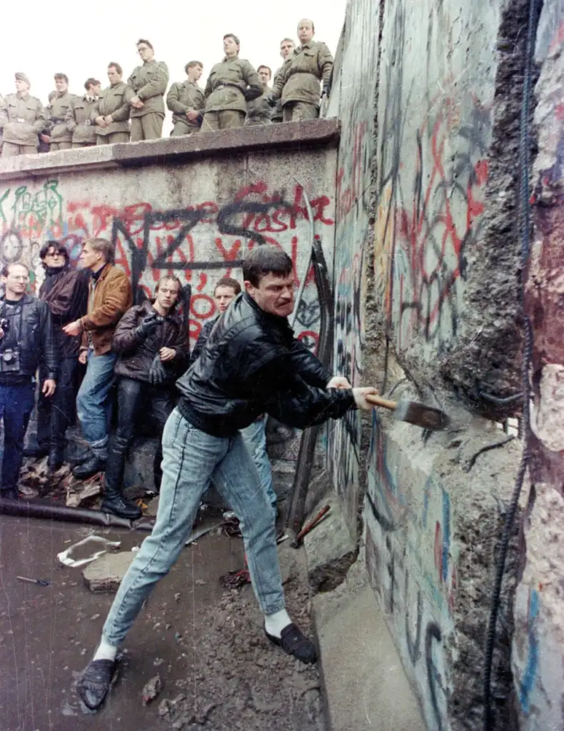 A man in a black jacket uses a pickaxe to chip away at a graffiti-covered wall. Several people watch, some holding cameras. Uniformed soldiers stand on top of the wall, observing the scene. The ground is wet with puddles.