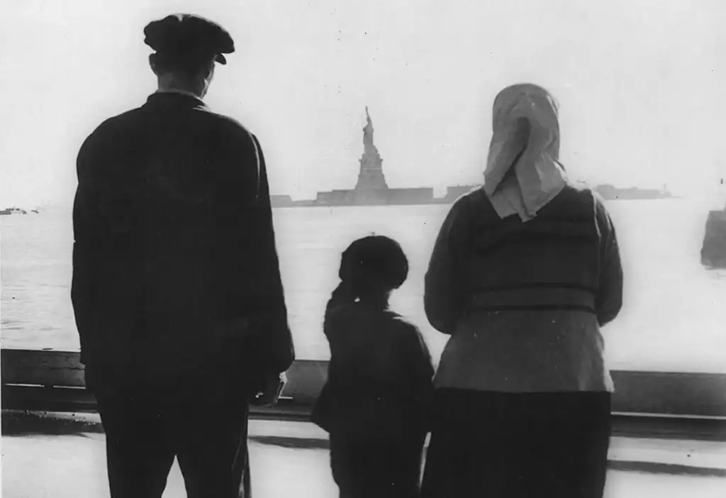 Three people stand on a boat, looking at the Statue of Liberty in the distance. The image is in black and white, depicting an adult wearing a cap, a child, and an adult with a headscarf, symbolizing immigration and hope.