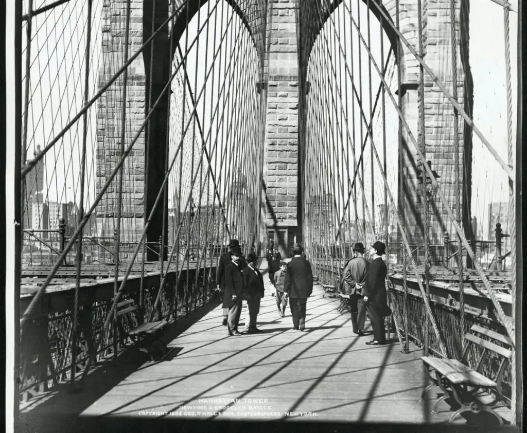 Black and white photo of people walking on the Brooklyn Bridge. Cables create a web-like pattern overhead. The city skyline is visible in the background.