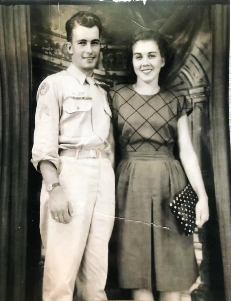 A black and white photo of a young man in a military uniform standing next to a woman in a patterned blouse and skirt. They are both smiling at the camera, standing against a backdrop with curtains. The woman holds a clutch.