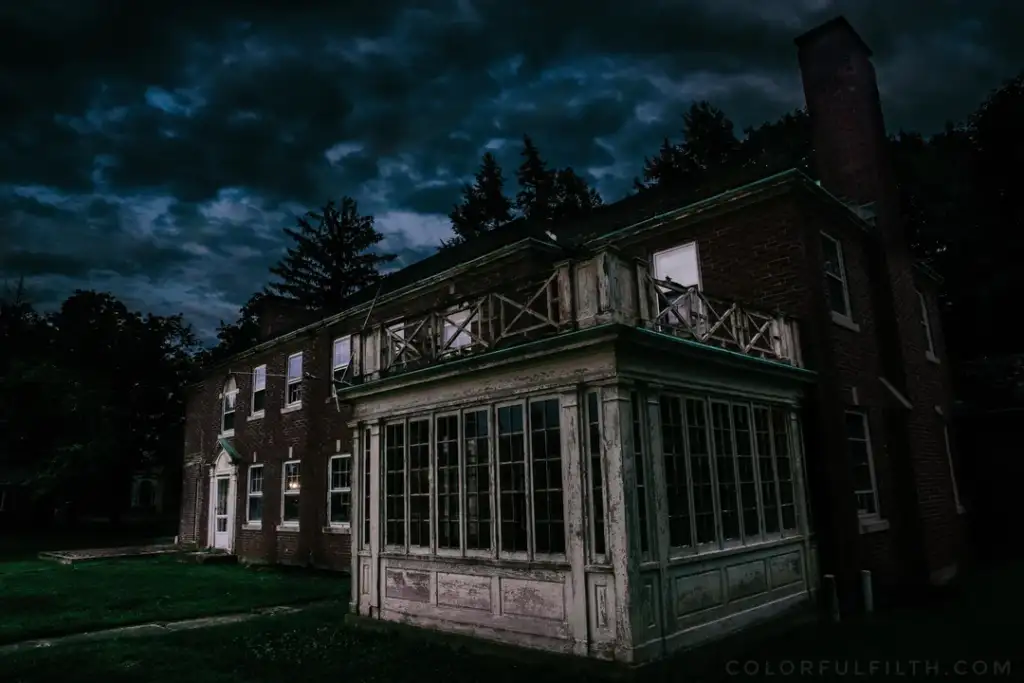 A dark, abandoned brick house with broken windows stands under an ominous, cloudy night sky. The building has a weathered appearance, with peeling paint and overgrown grass surrounding it. Shadows add a mysterious and eerie atmosphere.