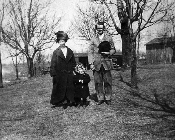 Vintage black and white photo of a family outdoors in winter clothing. A woman in a long coat and hat, a child in a hooded coat, and a man in a suit holding a hat stand between leafless trees, with a barn in the background.