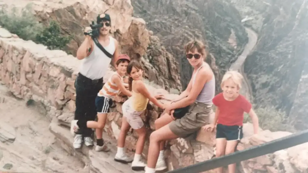 A group of five people, including four children and one adult with a video camera, sit on a rocky ledge overlooking a deep canyon. They're dressed casually in summer clothes, and the canyon walls create a dramatic backdrop.