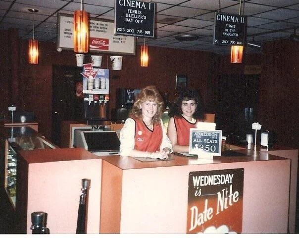 Two smiling employees in a vintage cinema lobby stand behind the concession counter. A sign on the counter reads "Admission All Seats $2.50." Above them, a marquee lists movie showtimes for "Ferris Bueller's Day Off" and "Top Gun.