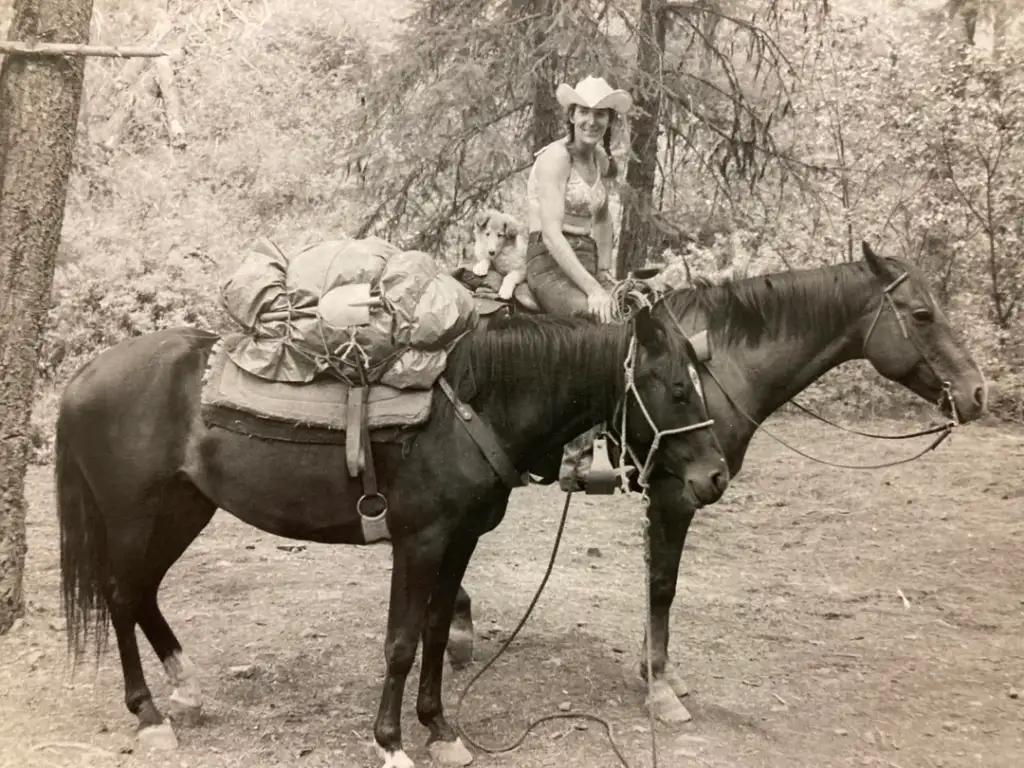 A woman wearing a cowboy hat sits on a horse in a forested area. The horse is heavily laden with gear. There is a dog sitting on top of the gear. The photo is in black and white.