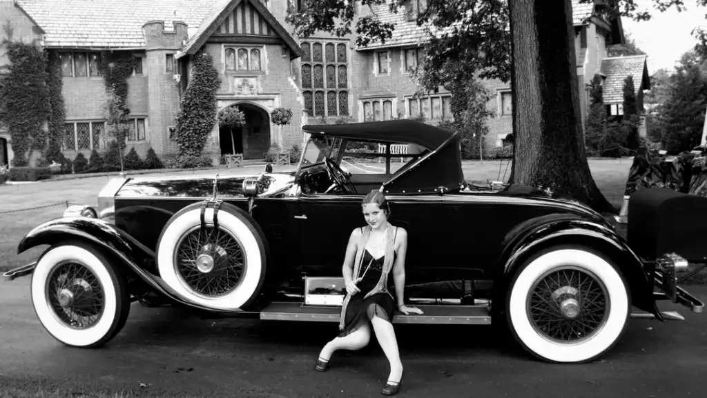 A black-and-white image of a woman in 1920s attire sitting on the running board of a vintage car with whitewall tires, parked in front of a large, ivy-covered mansion. The scene exudes a classic, elegant atmosphere.