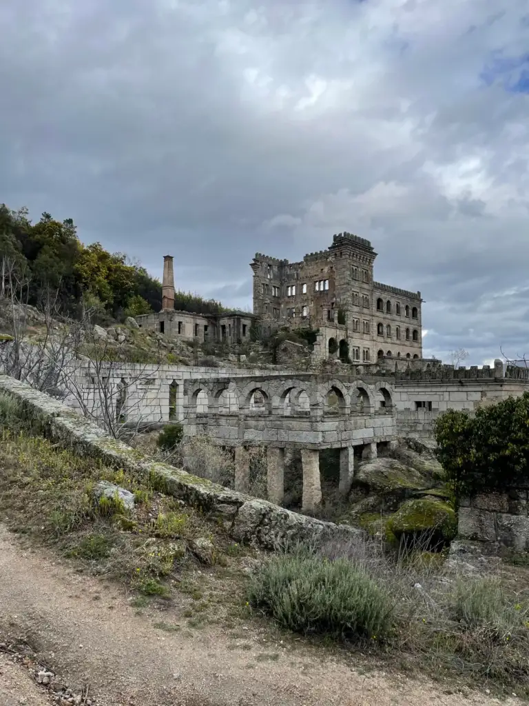 A historic, partially ruined stone building stands on a hillside under a cloudy sky. The structure features arches and ornate windows, with greenery and a dirt path in the foreground. Sparse trees grow around the building.