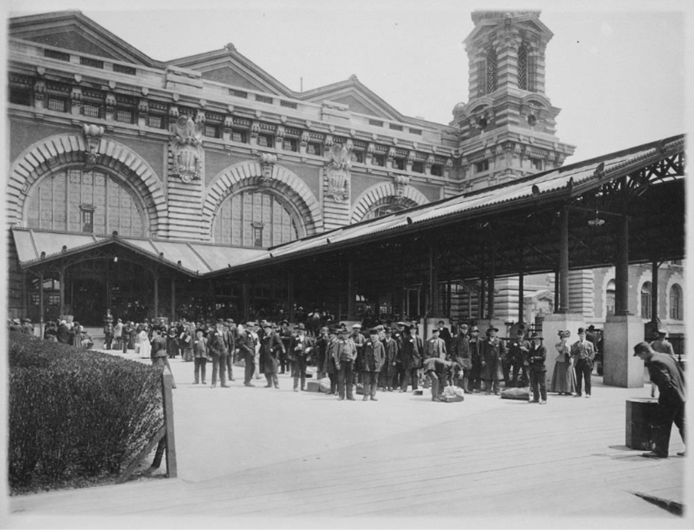 A historic black-and-white photograph shows a large crowd of people gathered outside a grand building with arched windows and ornate detailing. The scene captures diverse individuals, some with luggage, in a bustling outdoor setting.