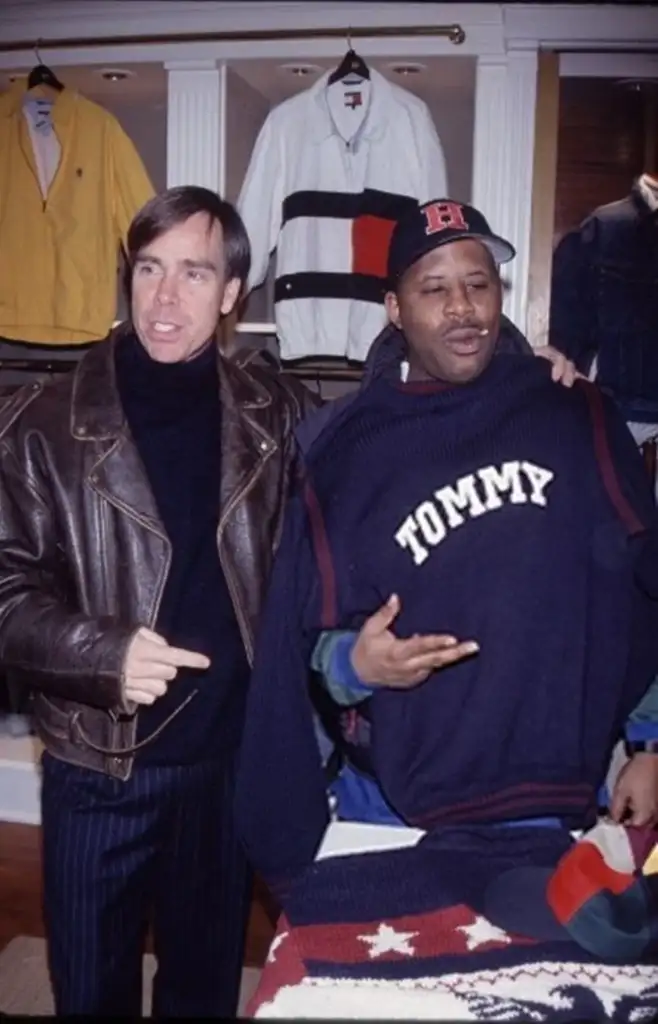 Two men in a clothing store. One wears a leather jacket and points at the other, who holds a navy Tommy Hilfiger sweater. The background displays various clothing items on shelves.