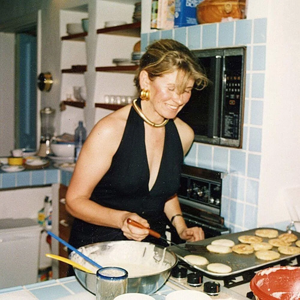 A woman in a sleeveless black dress is cooking in a kitchen. She is making pancakes or similar items on a stovetop. The kitchen has blue and white tiles and various cooking utensils are visible.