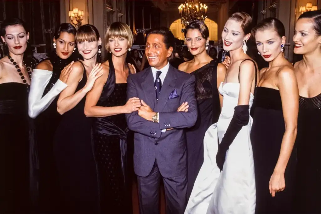 A well-dressed group of models in elegant evening gowns stand around a man in a suit. They are posing in a sophisticated indoor setting with ornate chandeliers and mirrors. The women display a variety of black and white outfit styles.