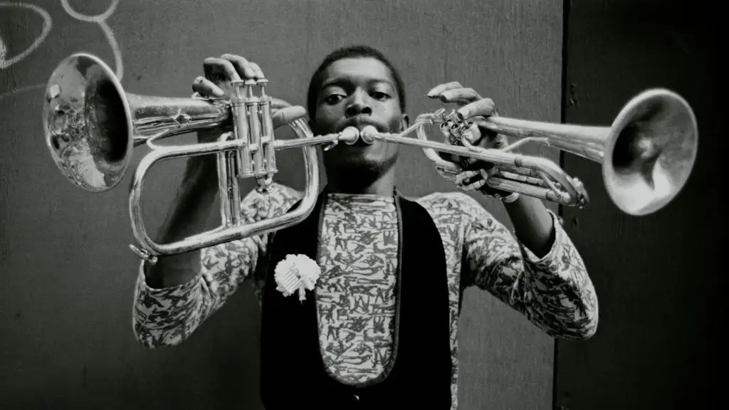 A person plays two trumpets simultaneously. They are wearing a patterned shirt and a vest adorned with a flower. The background is plain and dark, highlighting the unique performance.