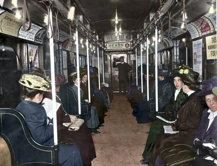 Vintage image of people sitting inside an early 20th-century subway car. Women and men wear formal clothing and hats. The carriage is decorated with ads and a conductor stands in the aisle. The atmosphere appears calm and composed.