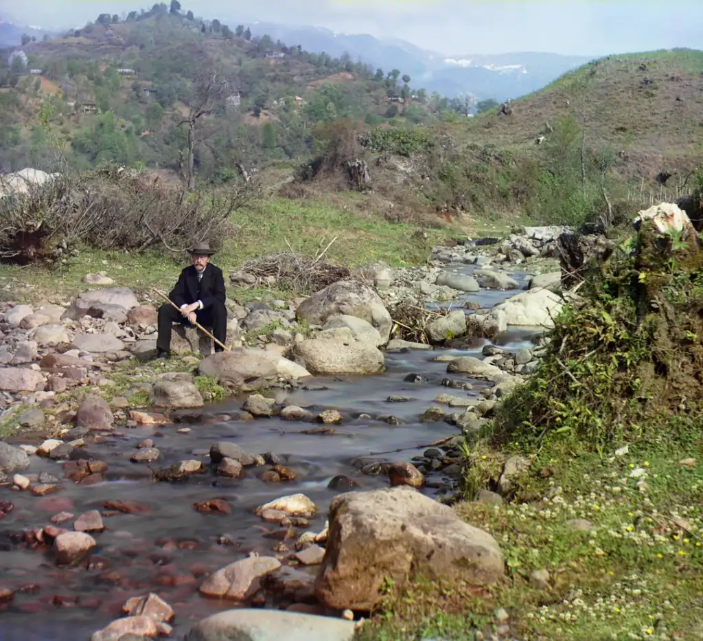 A man in a black outfit and hat sits on a rock beside a small, rocky stream. The stream flows through a grassy, hilly landscape with scattered trees and shrubs. Mountains are visible in the background under a partly cloudy sky.