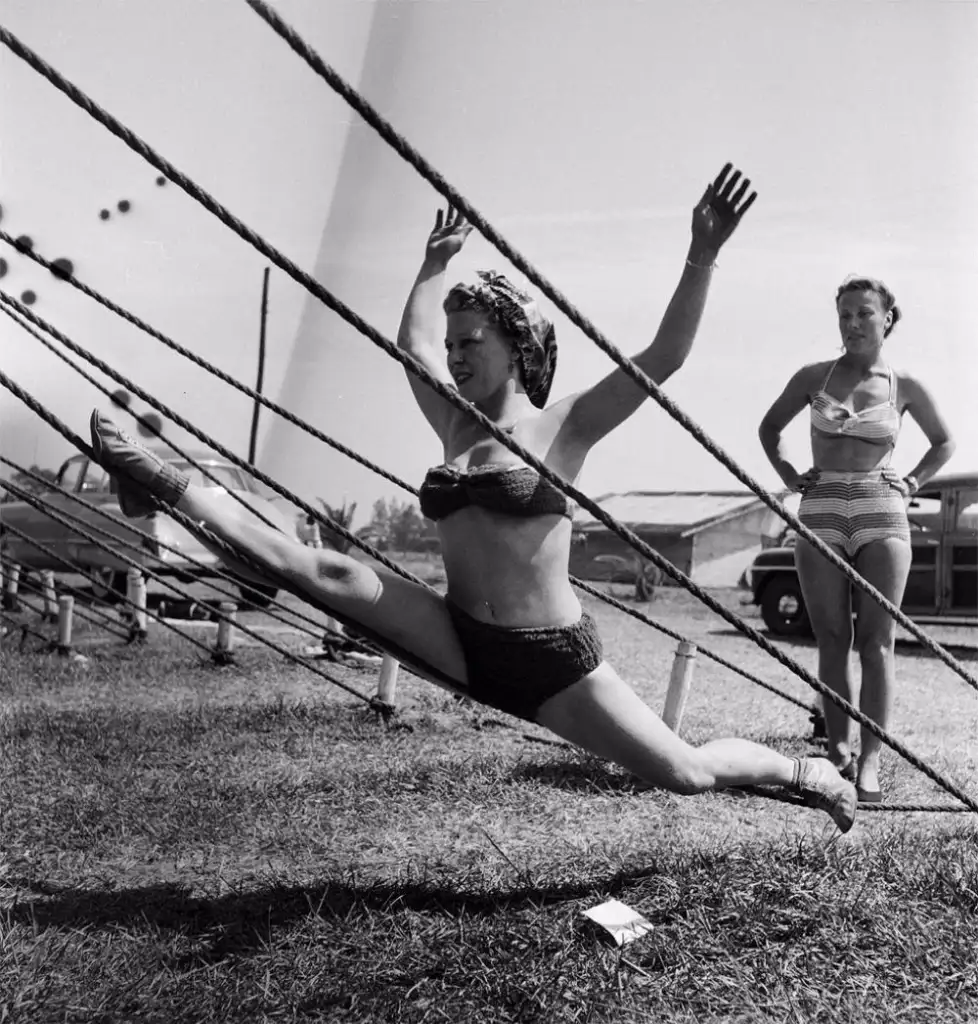 A woman in a bikini performs a split between two tensioned ropes, with arms raised for balance. Another woman in a striped bikini stands nearby with hands on hips, watching. Both are outdoors on a grassy area with vehicles and structures in the background.