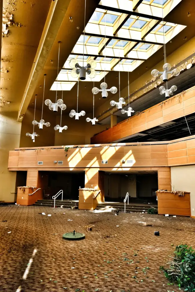 Abandoned spacious room with a high ceiling featuring a grid of hanging light bulbs. The worn carpet and scattered debris cover the floor. Sunlight streams through the large skylight, illuminating the wooden panels and overall decay.