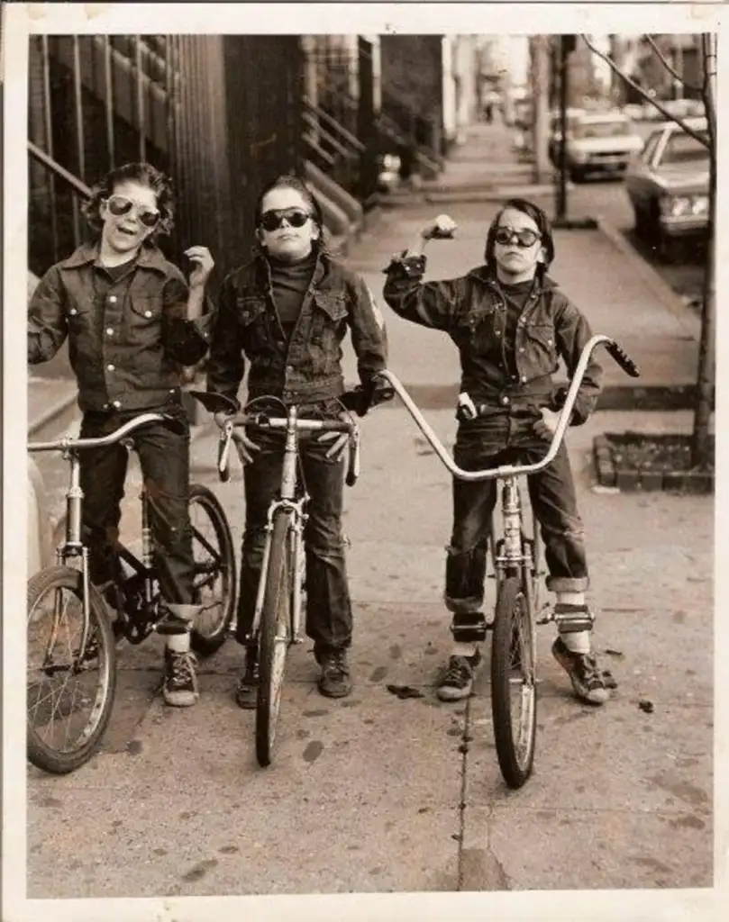 Three kids wearing sunglasses ride bicycles on a city street. They are dressed in denim and appear playful and confident, striking cool poses for the photo. The background features buildings and parked cars.