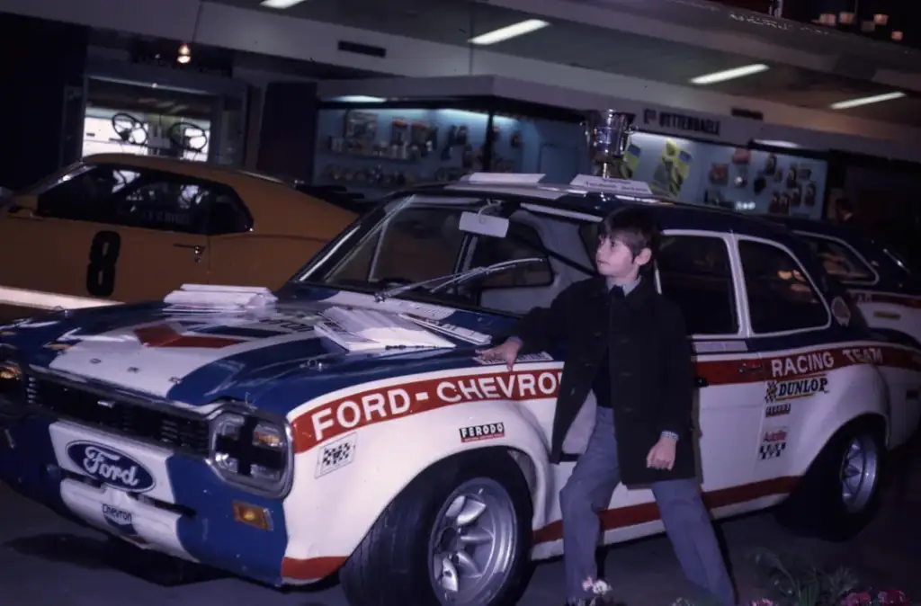 A young boy leans against a classic Ford racing car with "Ford" and "Chevron" branding. The car has a blue and white paint job with red accents and is displayed indoors, surrounded by other vehicles.